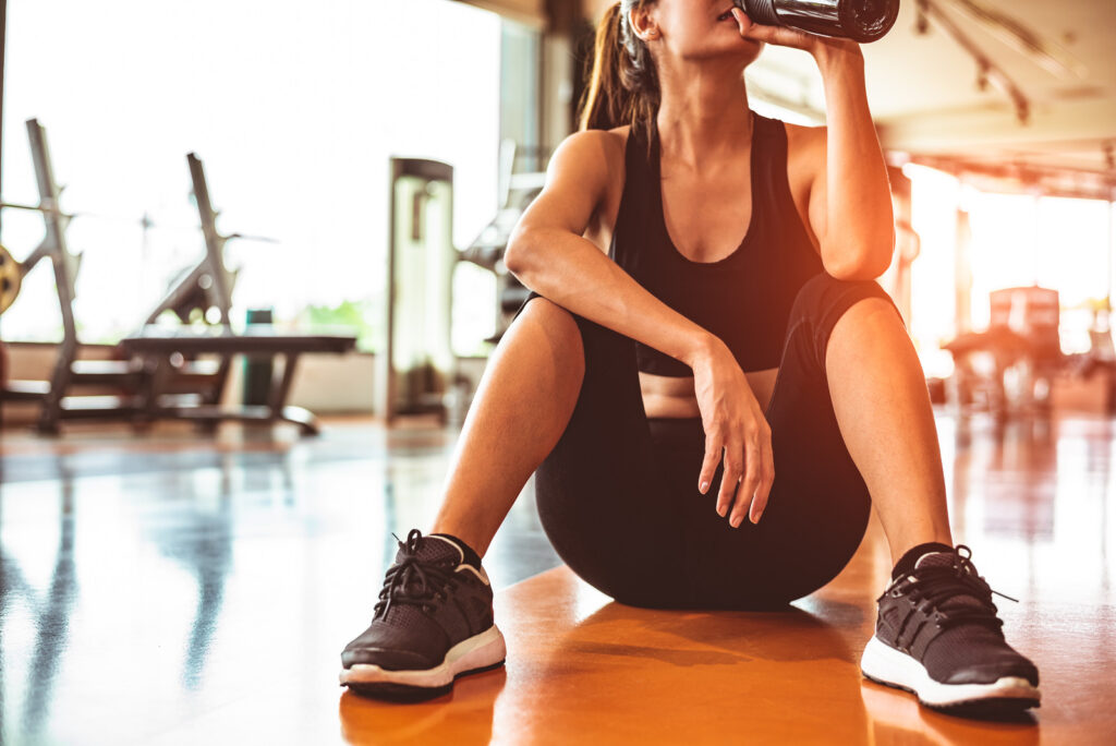 Sport woman relax resting after workout or exercise in fitness gym. Sitting and drinking protein shake or drinking water on floor. Strength training and Bodybuilder muscle theme. Warm and cool tone - FitnessLife Magazine